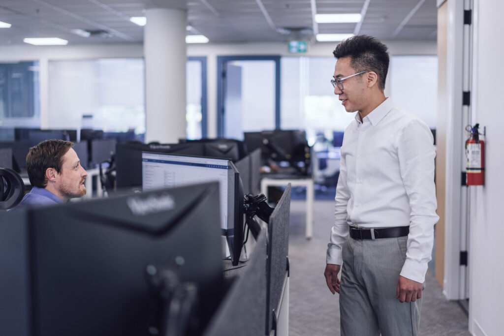 A coworker standing talking to another sitting at their desk